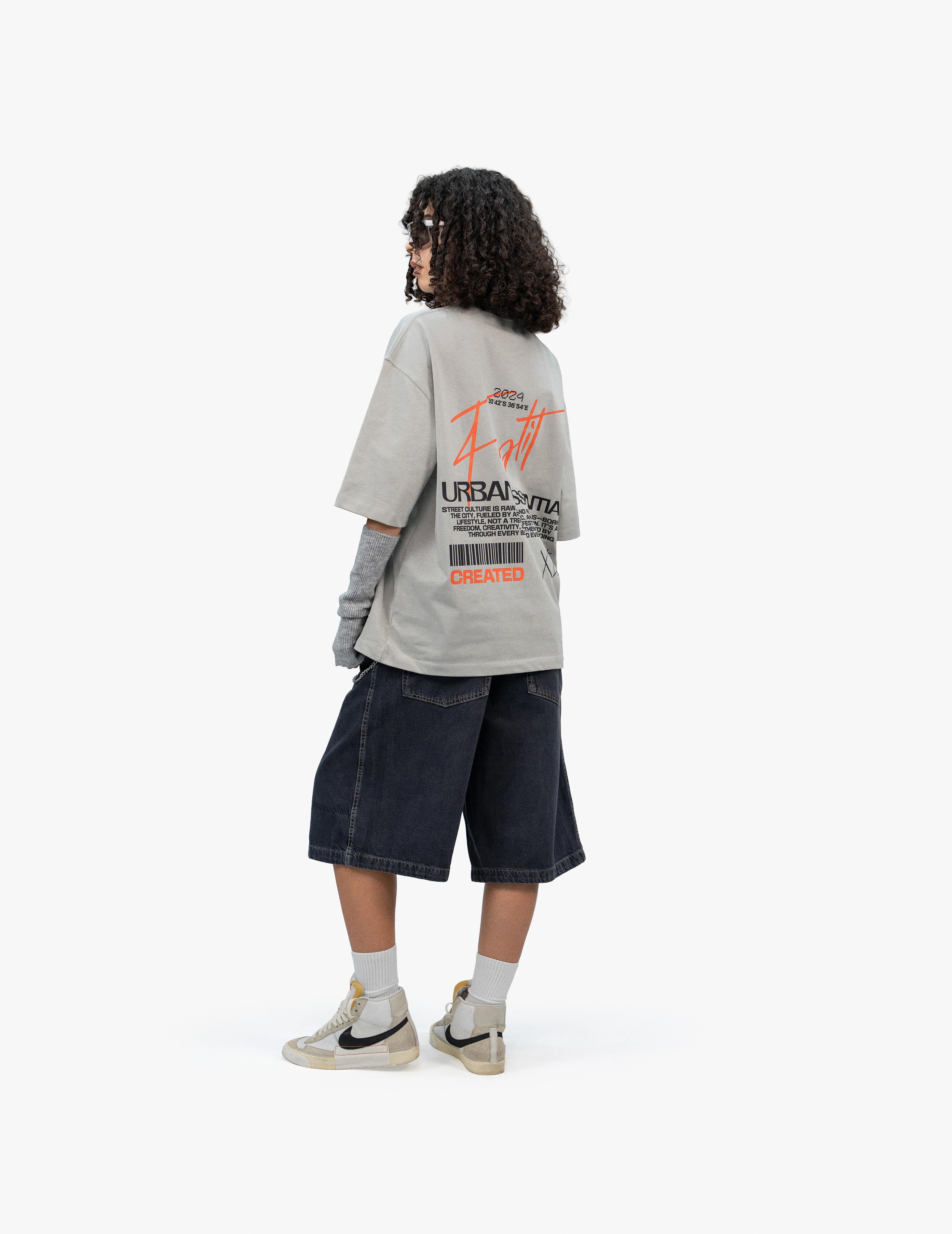 Urban Essential Limited Edition Gri Oversize Tişört,  Beyaz bir stüdyo fonunun önünde, arkası dönük ve hafifçe sağa dönük bir şekilde duran, kısa siyah kıvırcık saçlı bir kadın. Üzerinde sırt kısmında turuncu ve siyah renklerde, barkod ve yazılar içeren grafik baskılar bulunan açık gri, geniş kesim bir tişört ve lacivert, bol kesim, diz hizasında bir şort giyiyor. Tişörtün altında gri renkli kol ısıtıcıları görülüyor. Ayaklarında bilekli spor ayakkabılar ve beyaz çoraplar bulunuyor. -  Faylit