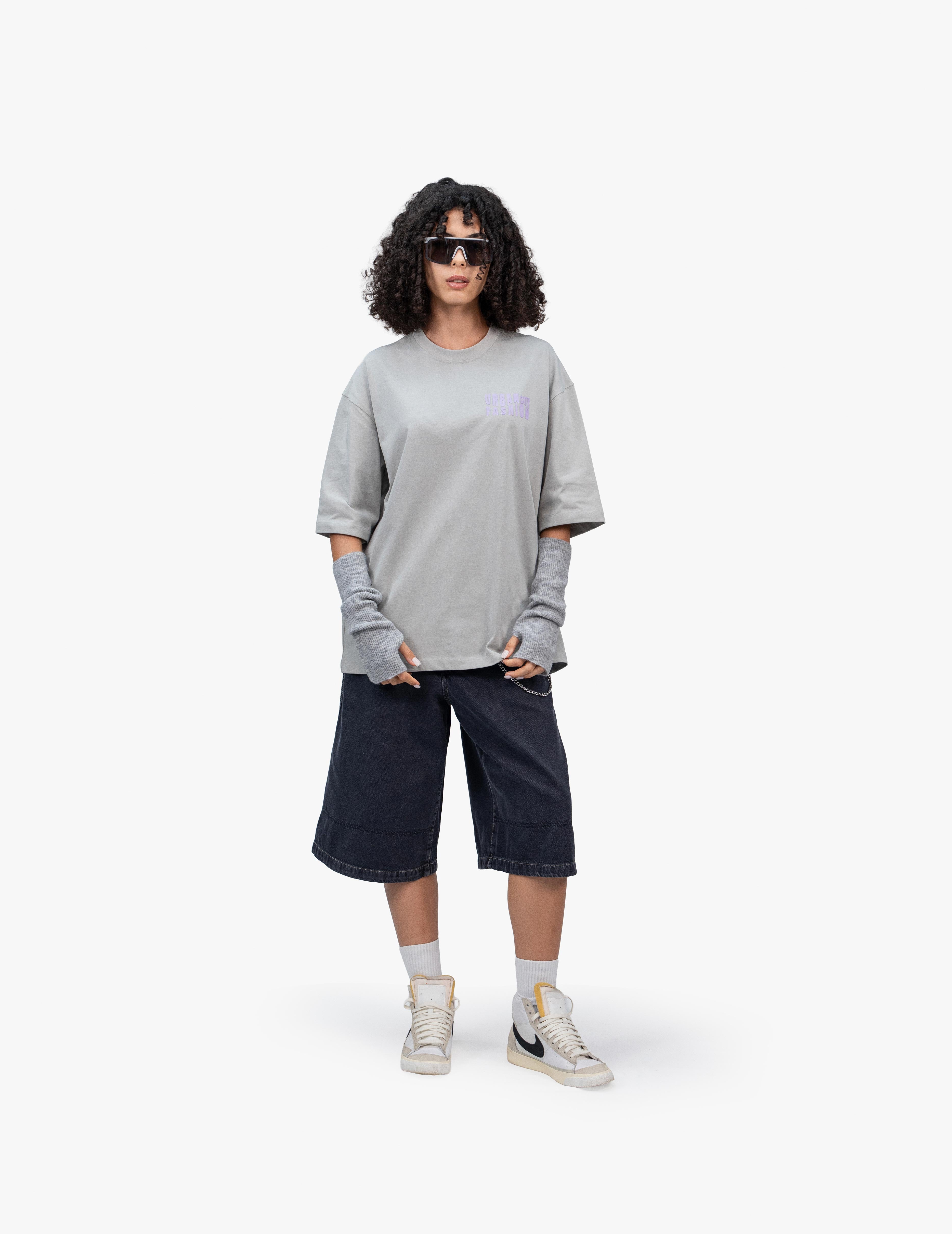 Urban Essential Limited Edition Gri Oversize Tişört,  Beyaz bir stüdyo fonunun önünde, doğrudan kameraya bakan, kısa siyah kıvırcık saçlı bir kadın. Beyaz çerçeveli futuristik görünümlü bir güneş gözlüğü takıyor ve ellerini tişörtünün cebine sokmuş. Üzerinde açık gri, geniş kesim bir tişört ve lacivert, bol kesim, diz hizasında bir şort giyiyor. Tişörtün altında gri renkli kol ısıtıcıları görülüyor. Ayaklarında bilekli spor ayakkabılar ve beyaz çoraplar bulunuyor. -  Faylit