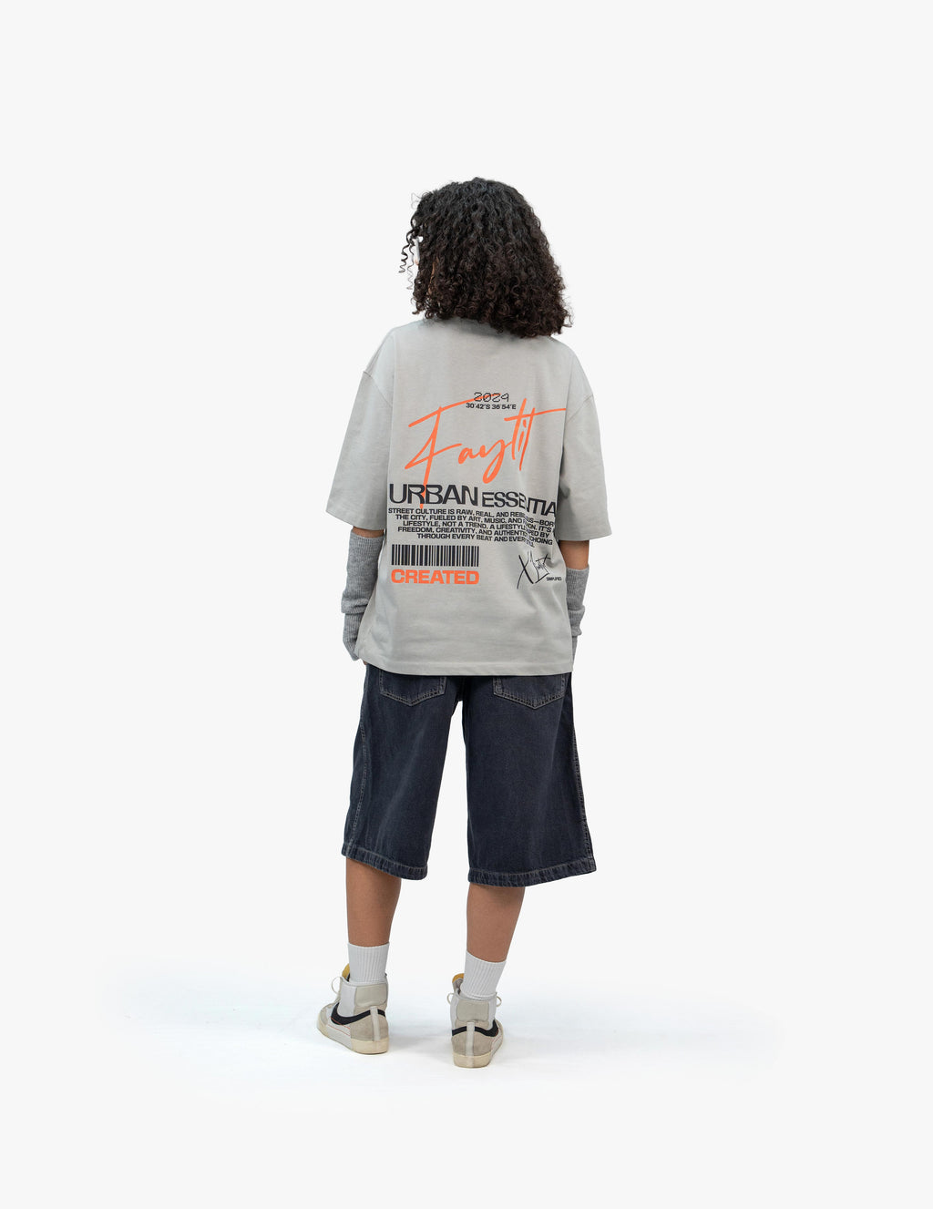 Urban Essential Limited Edition Gri Oversize Tişört,  Beyaz bir stüdyo fonunun önünde, arkası dönük ve hafifçe sola dönük bir şekilde duran, kısa siyah kıvırcık saçlı bir kadın. Üzerinde sırt kısmında turuncu ve siyah renklerde, barkod ve yazılar içeren grafik baskılar bulunan açık gri, geniş kesim bir tişört ve lacivert, bol kesim, diz hizasında bir şort giyiyor. Tişörtün altında gri renkli kol ısıtıcıları görülüyor. Ayaklarında bilekli spor ayakkabılar ve beyaz çoraplar bulunuyor. -  Faylit