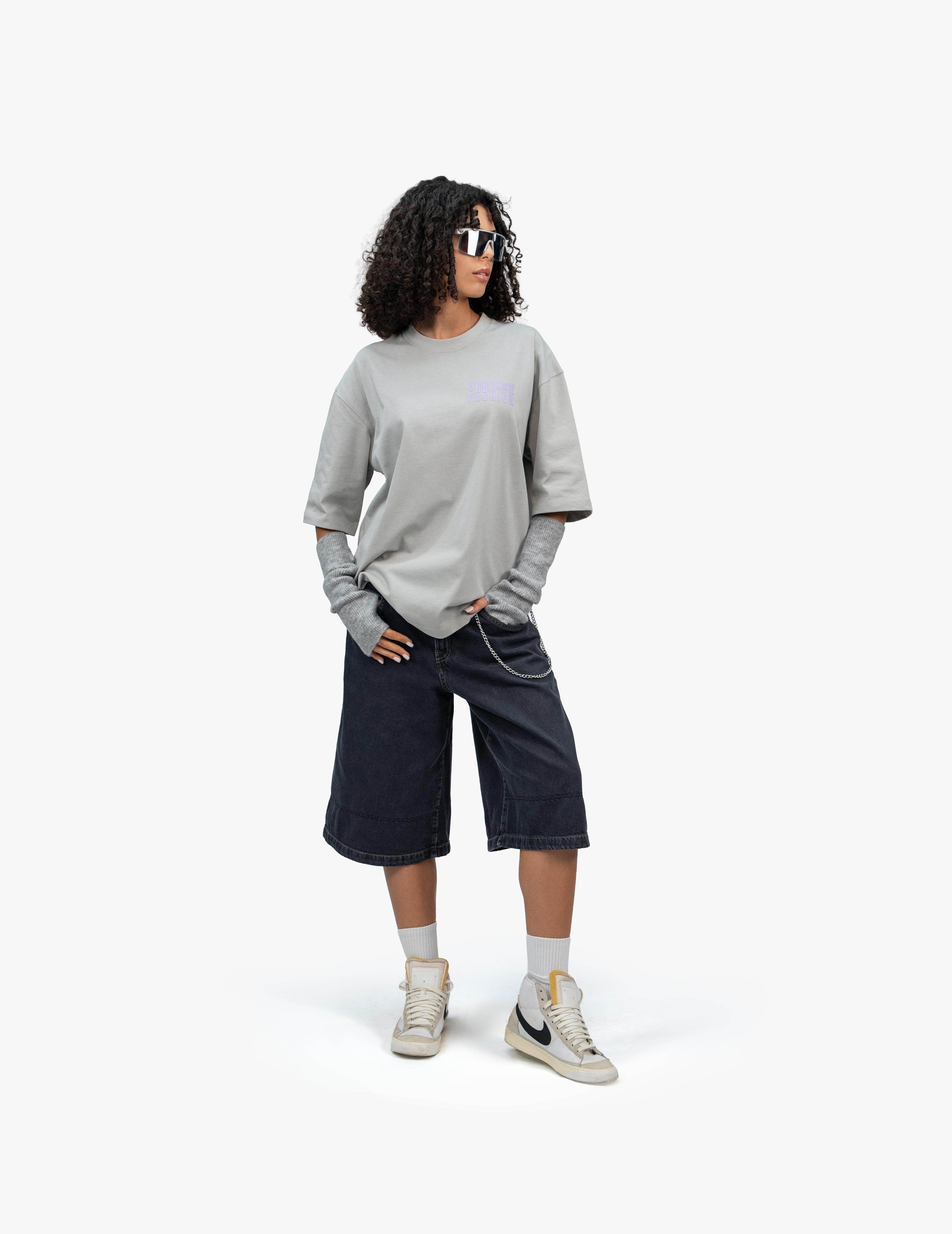 Urban Essential Limited Edition Gri Oversize Tişört, Beyaz bir stüdyo fonunun önünde, hafifçe sağa dönük duran, kısa siyah kıvırcık saçlı bir kadın. Beyaz çerçeveli futuristik görünümlü bir güneş gözlüğü takıyor. Üzerinde açık gri, geniş kesim bir tişört ve lacivert, bol kesim, diz hizasında bir şort giyiyor. Tişörtün altında gri renkli kol ısıtıcıları ve sağ bacağından sarkan zincir detayı görülüyor. Ayaklarında bilekli spor ayakkabılar ve beyaz çoraplar bulunuyor. -  Faylit