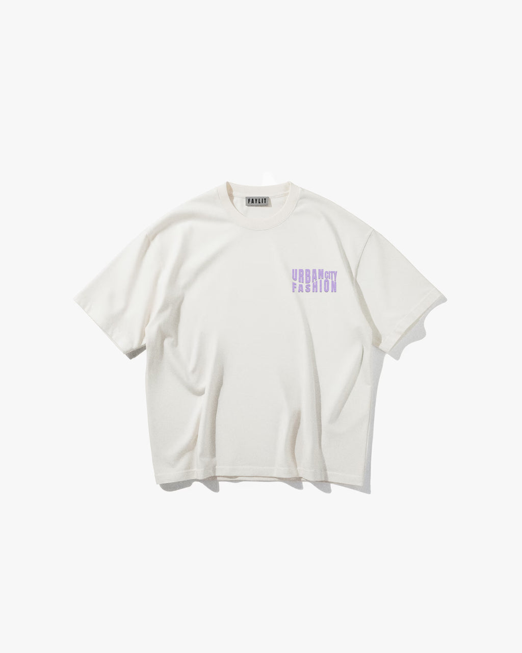 Urban Essential Limited Edition Off White Oversize Tişört - Ekru rengi oversize tişörtün ön yüzünün düz çekim ürün fotoğrafı. Tişörtün sol göğsünde, lavanta ve mor tonlarında, bloklu/pikselli bir fontla üç satır halinde "URBAN CITY FASHION" yazan bir logo bulunmaktadır. Yaka içindeki marka etiketi de görülebilir. -  Faylit

