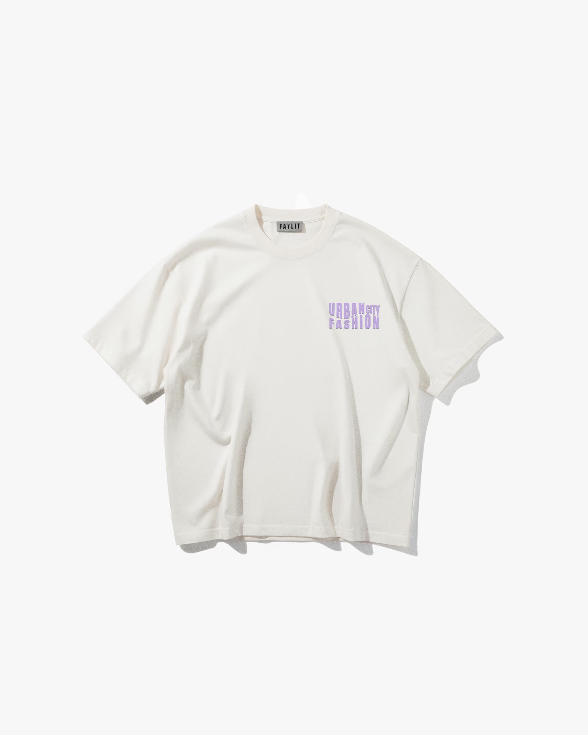Urban Essential Limited Edition Off White Oversize Tişört - Ekru rengi oversize tişörtün ön yüzünün düz çekim ürün fotoğrafı. Tişörtün sol göğsünde, lavanta ve mor tonlarında, bloklu/pikselli bir fontla üç satır halinde "URBAN CITY FASHION" yazan bir logo bulunmaktadır. Yaka içindeki marka etiketi de görülebilir. -  Faylit
