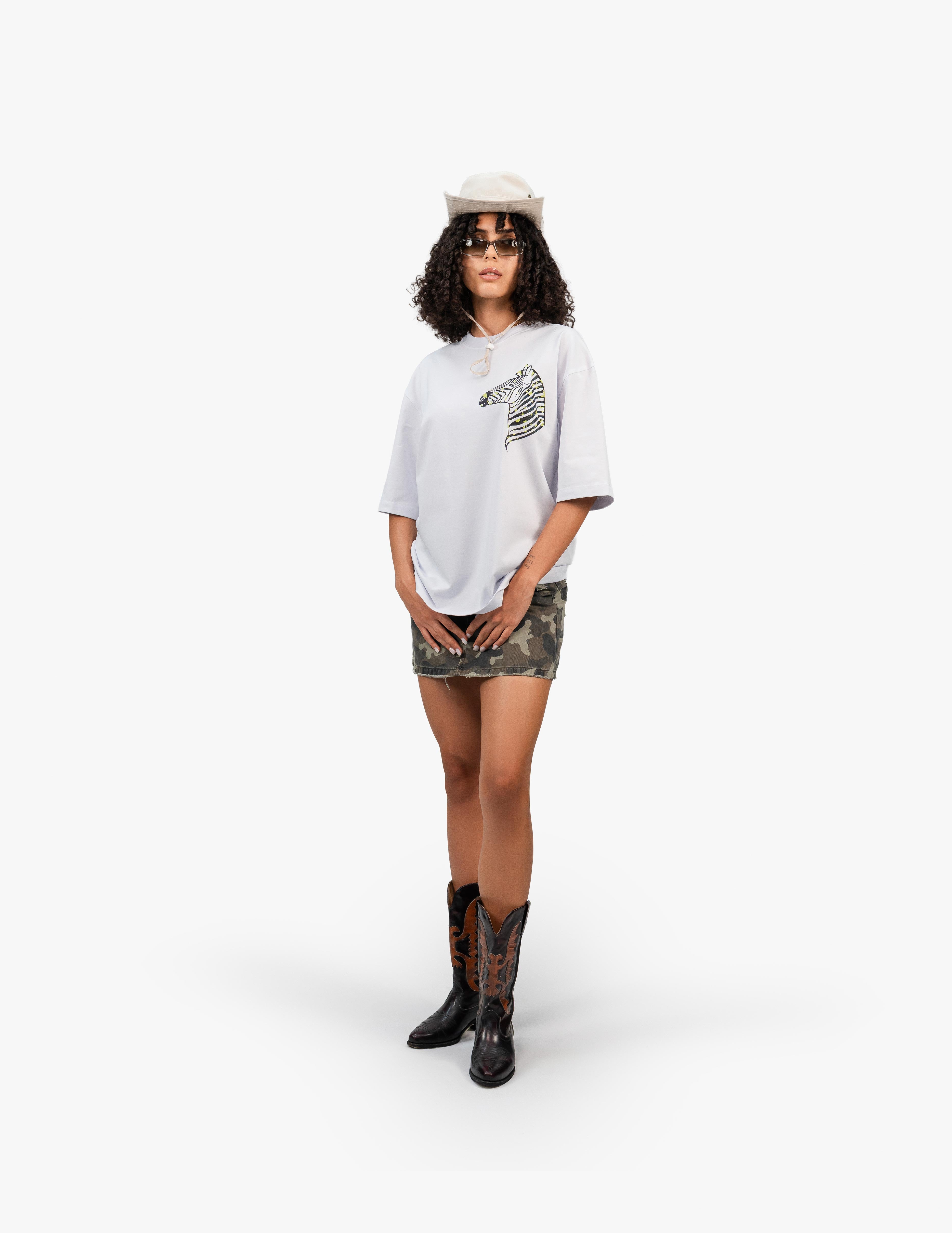 Zebra Holografik Flok Baskı Beyaz Oversize Tişört,  Beyaz bir stüdyo fonunun önünde, doğrudan kameraya bakan, kısa siyah kıvırcık saçlı bir kadın. Beyaz bir kovboy şapkası ve siyah çerçeveli güneş gözlüğü takıyor. Üzerinde göğüs kısmında zebra desenli bir grafik baskı bulunan açık gri, geniş kesim bir tişört ve kamuflaj desenli mini bir etek giyiyor. Ayaklarında siyah ve turuncu desenli kovboy çizmeleri bulunuyor. -  Faylit