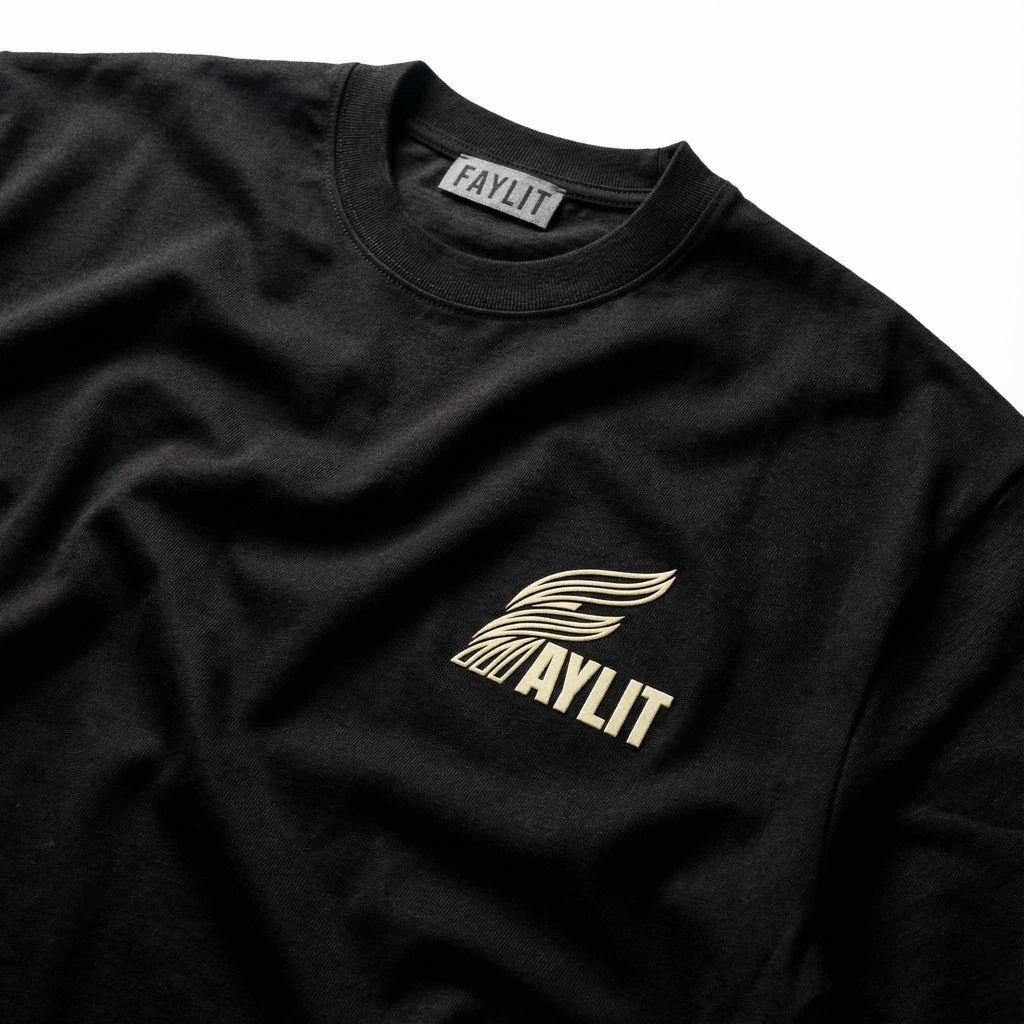 Siyah, yüksek gramajlı pamuklu kumaşın dokusunu ve kalitesini gösteren yakın çekim detay. Tişörtün sol göğsünde 'FAYLIT' yazısı ve üzerinde kanat ikonu bulunan, hafif kabartmalı (500 micron 3D) ve dokulu krem rengi logo işlemesi net bir şekilde görülüyor. Yaka kısmındaki fitilli ribana detayları ve iç kısımdaki gri 'FAYLIT' marka etiketi ürünün premium işçiliğini yansıtıyor.