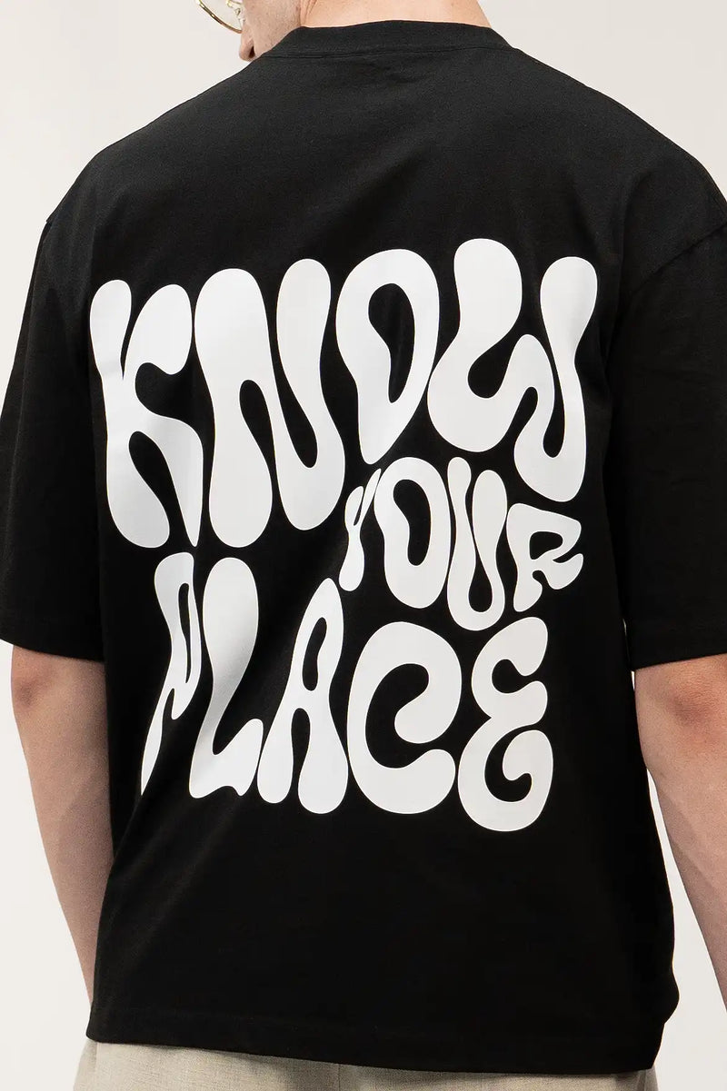 siyah oversize tişört giyen bir model. Siyah tişörtün arkasında 'KNOW YOUR PLACE' motto yazısı dikkat çekiyor.