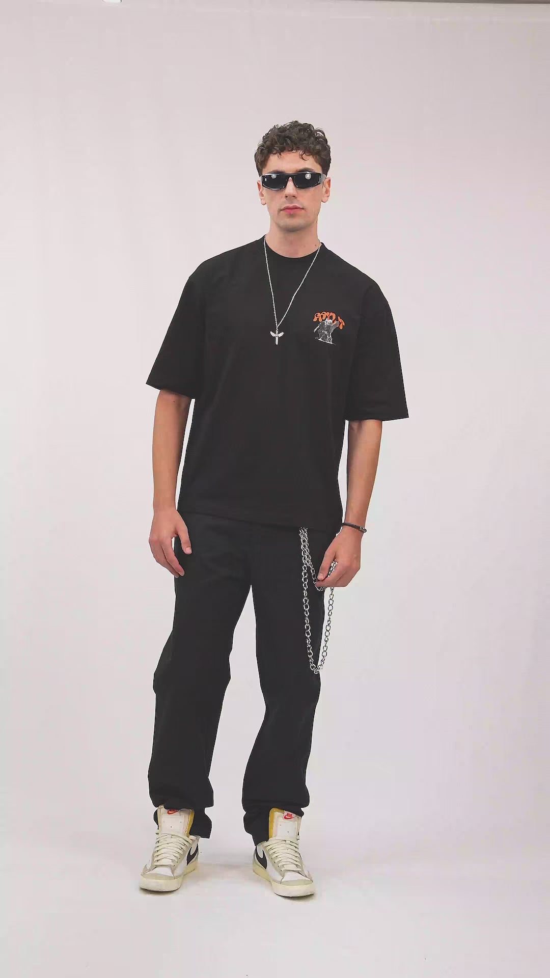 Faylit Persistence Retro Grafik siyah oversize tişört giyen güneş gözlüklü bir foto model. Tişört tuğla turuncusu rengiyle dikkat çekiyor. Manken ayrıca melek tasarımlı özel bir kolye takıyor. Siyah kargo (cargo) pantolonunda bir kot zinciri bulunuyor ve son model nike sneaker giyiyor.