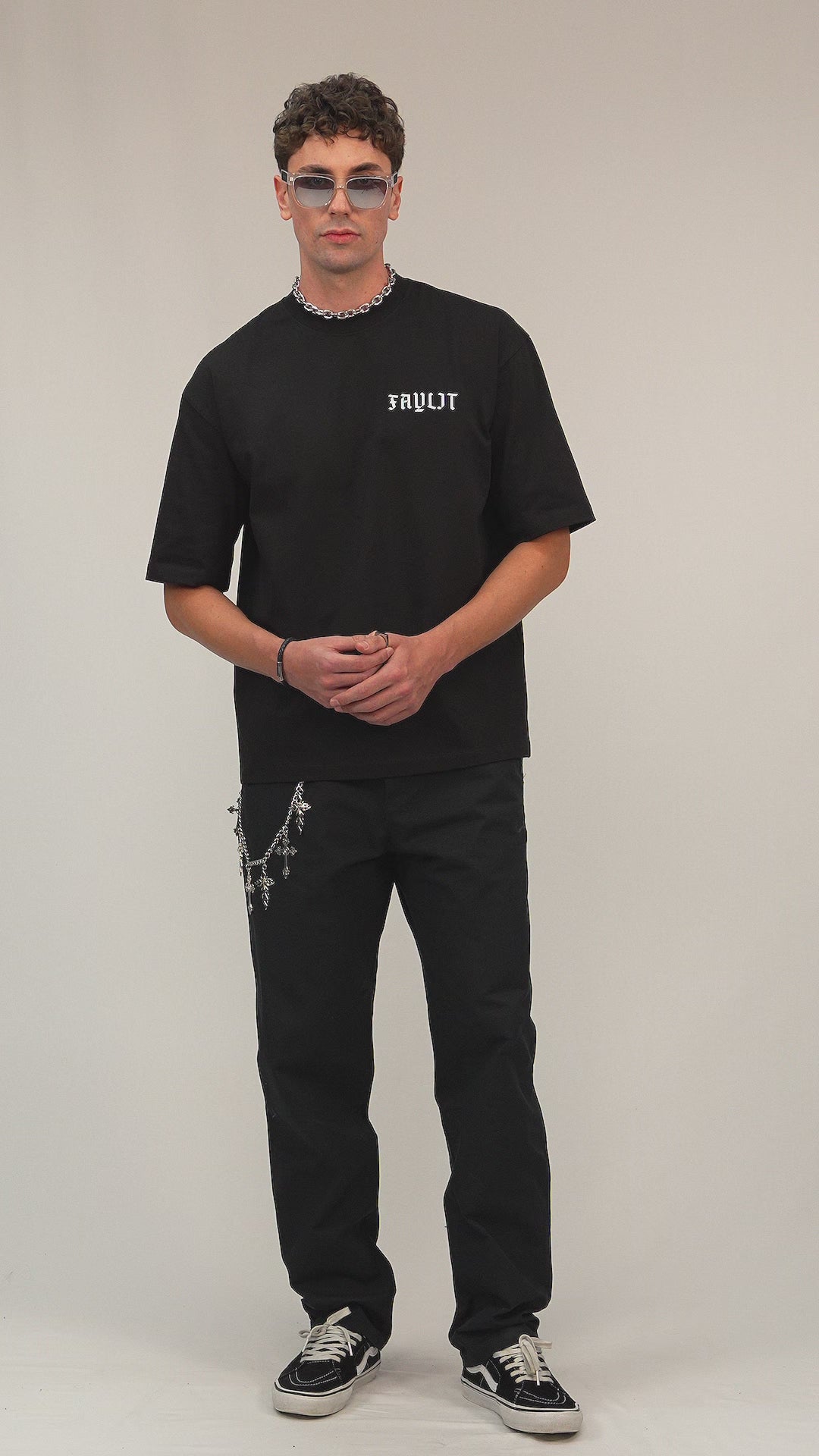 Faylit Arp Angels Siyah Premium oversize tişört giyen bir foto model. Aksesuar olarak gümüş renginde aynalı rayban gözlük takıyor. Kolye ve kot zinciri detayı dikkat çekiyor. Mankenin ayağında ise vans model ayakkabı mevcut