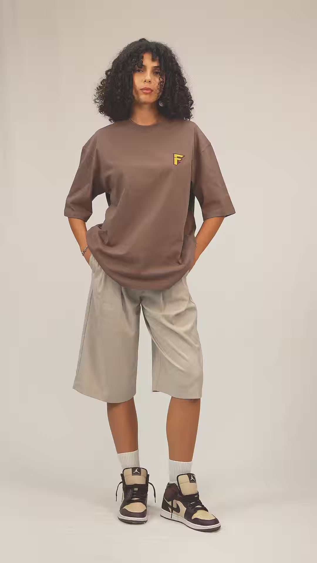 Faylit Kahverengi unisex oversize tişört giyen kıvırcık saçlı model. Kombini basketbol şortu ile tamamlamış ve ayakkabıları son model jordan.